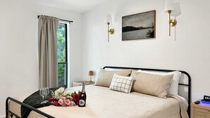 Ein ordentlich bezogenes Bett mit einem Blumenstrauß und einer Champagnerflasche, ein Bild an der Wand und ein Fenster mit Vorhängen.