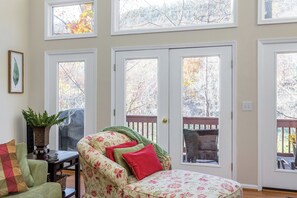 A room with a green armchair, a small table, and a potted plant. There are three doors with windows, and a view of trees outside.