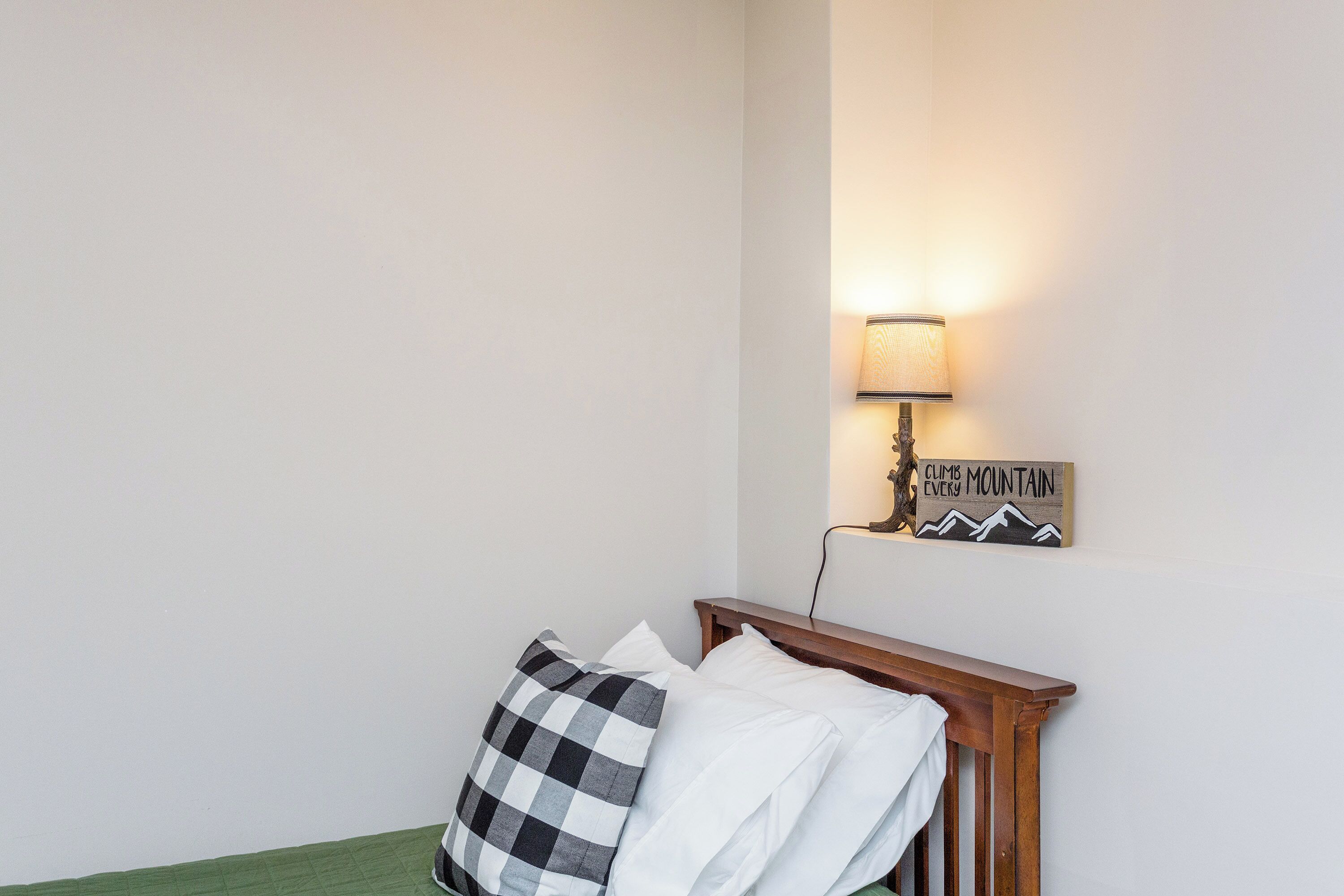 A bedroom with a wooden headboard, a green bedspread, a checkered pillow, a white pillow, a wall-mounted lamp, and a motivational sign.