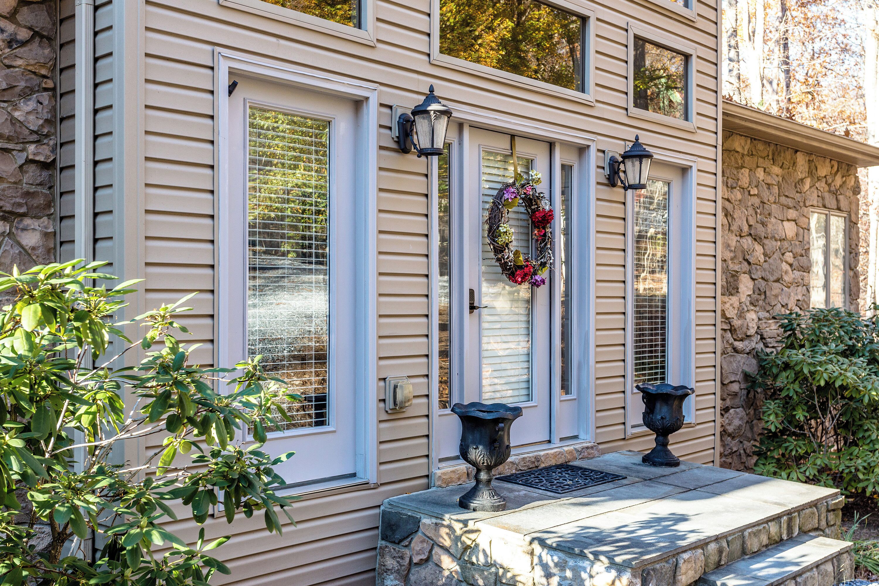 A house with beige siding, white trim, and a stone foundation. There are three windows with white shutters, a decorative wreath on the door, and two wall-mounted lanterns.