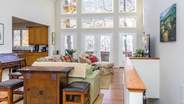 A spacious living room with a high ceiling, large windows, a dining area, and a kitchen.