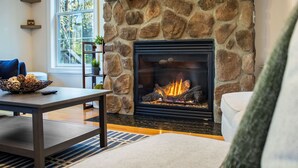 A stone fireplace with a fire, a wooden table with a bowl of bread, and a couch in a living room with a large window. - Sunrise Views & Mountain Charm! (Mc Gaheysville)