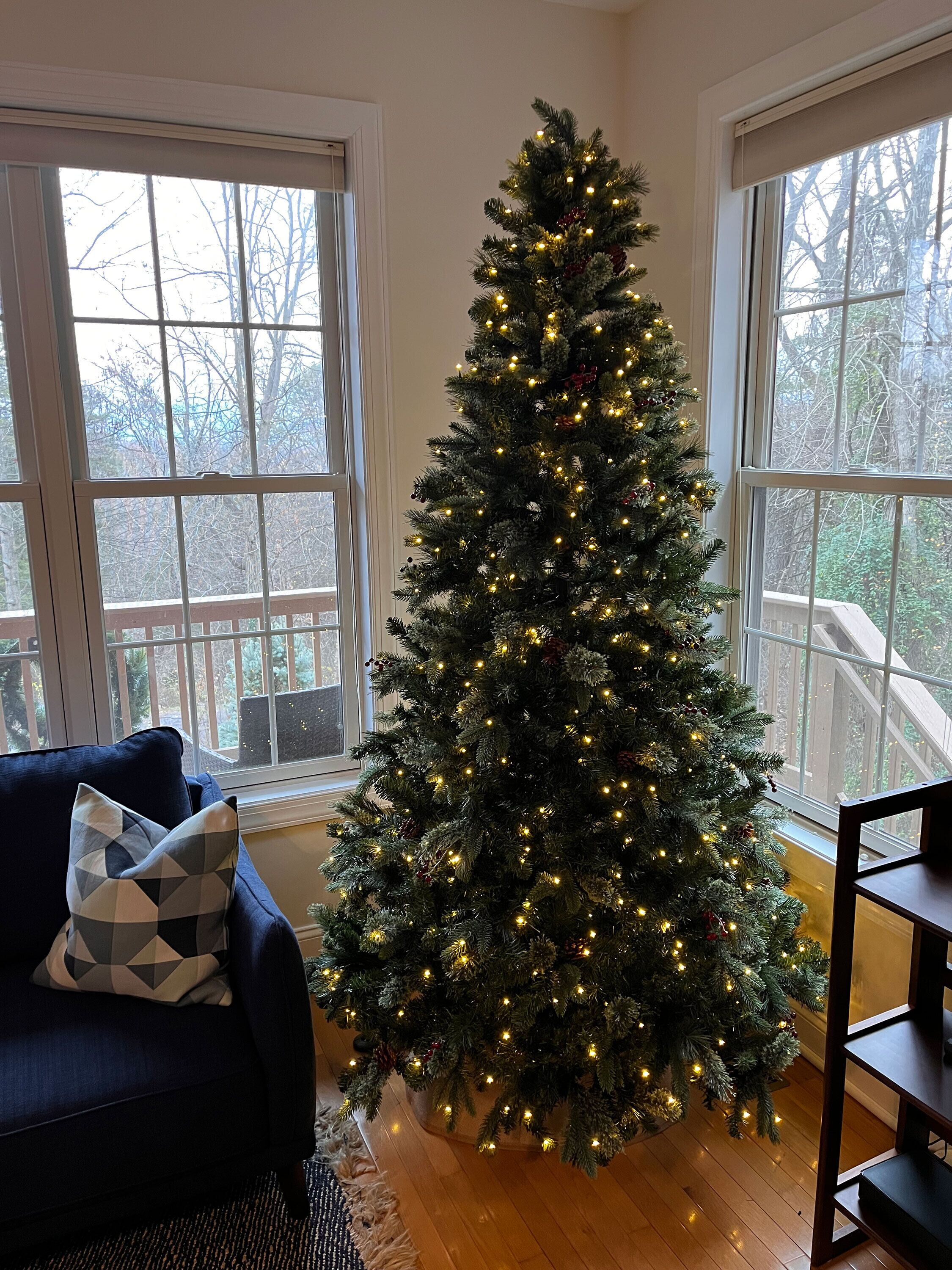 Een kerstboom in een kamer met grote ramen, een donkerblauwe bank en een houten vloer.