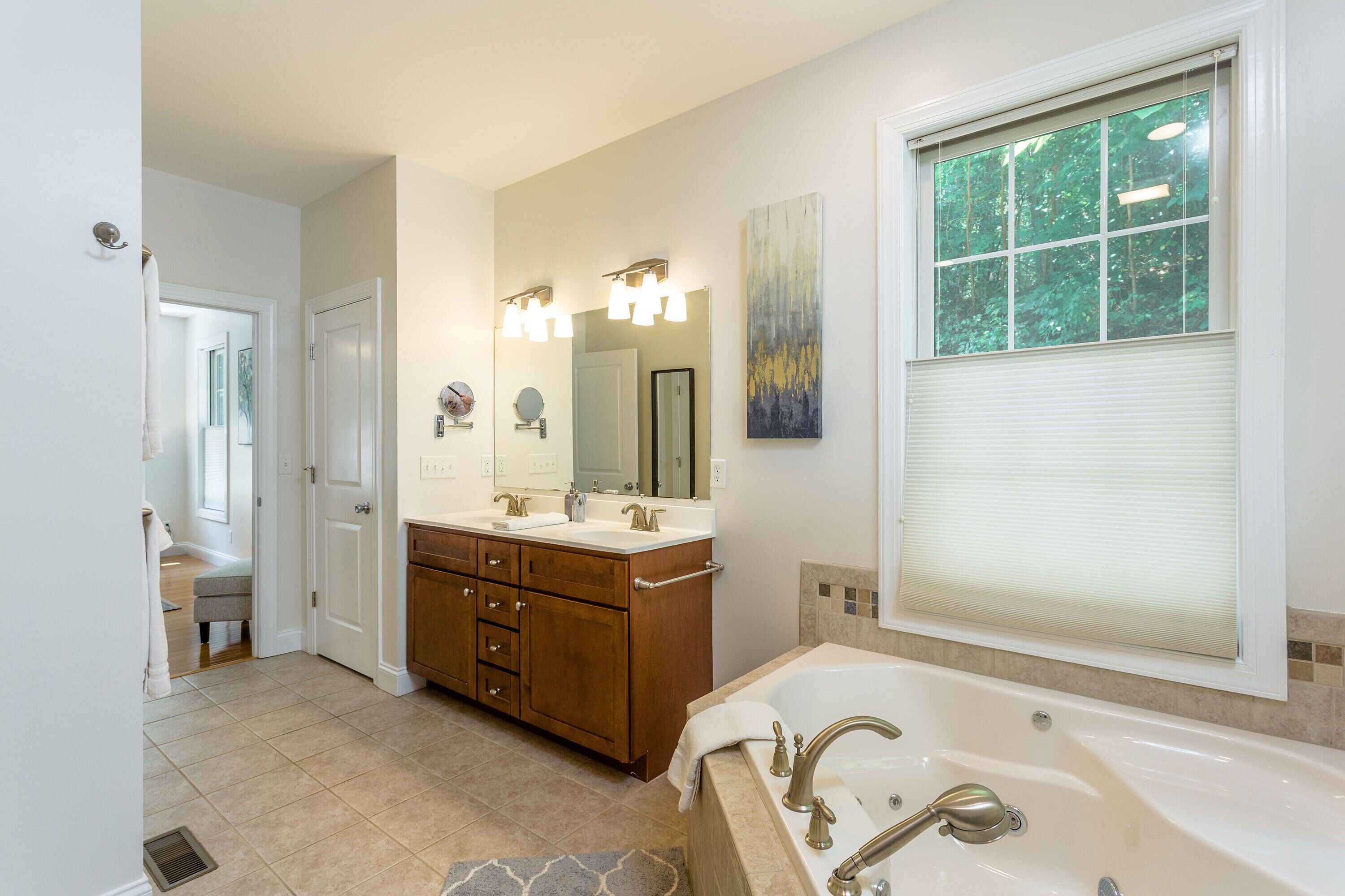 A modern bathroom with a large bathtub, double vanity with a mirror, and a window with blinds.
