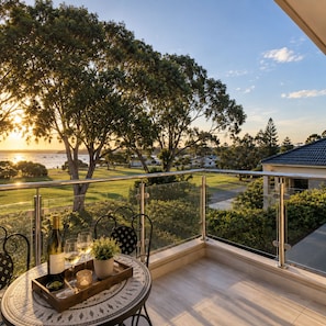 A balcony with a table set for a meal, overlooking a scenic view of a golf course and ocean during sunset.