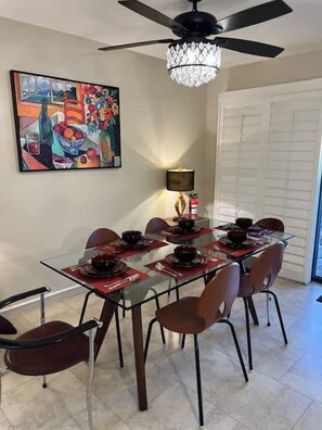 A dining area with a glass table, red placemats, and black bowls. There is a chandelier, a painting on the wall, and a door with blinds.