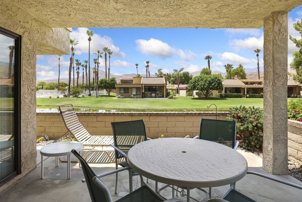 A covered patio with a round table and chairs, overlooking a well-maintained lawn and buildings in the background. - Spacious Single-story Condo With Golf Course Views (Palm Springs)
