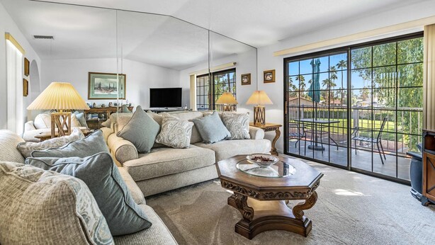 A spacious living room with a large sectional sofa, a glass coffee table, and a television. The room has large windows offering a view of the outdoors. - Stylish Condo With Panoramic View Community Pool (Palm Springs)