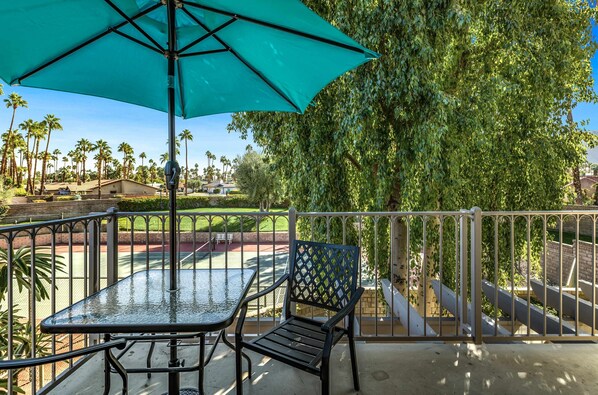 A balcony with a table and chairs, a turquoise umbrella, and a view of a golf course and palm trees. - Stylish Condo With Panoramic View Community Pool (Palm Springs)