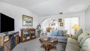 A living room with a large flat-screen TV, a wooden entertainment unit, a glass coffee table, a grey sofa, and a dining area with a wooden table and chairs. - Stylish Condo With Panoramic View Community Pool (Palm Springs)