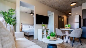 Uma sala de estar moderna com sofá, mesa de centro redonda e televisão na parede. Há uma área de jantar com mesa e cadeiras, e uma cozinha com geladeira.