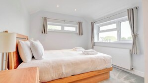 A bedroom with a wooden bed, white linens, a bedside table with a lamp, and a window with curtains.
