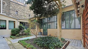 A courtyard with a brick pathway, a small garden area, and a building with large windows and a stone facade.