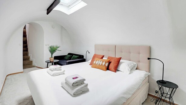 A modern bedroom with a large bed, a small table, a chair, a potted plant, and a staircase leading to an upper level.