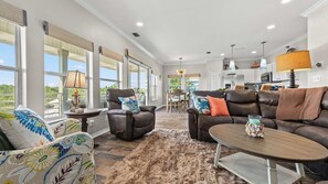 A spacious living room with a large window, leather sofas, a dining area, and a kitchen in the background.