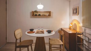 A dining area with a round table, two chairs, a hanging light fixture, a shelf with decorative items, and a desk with a lamp.