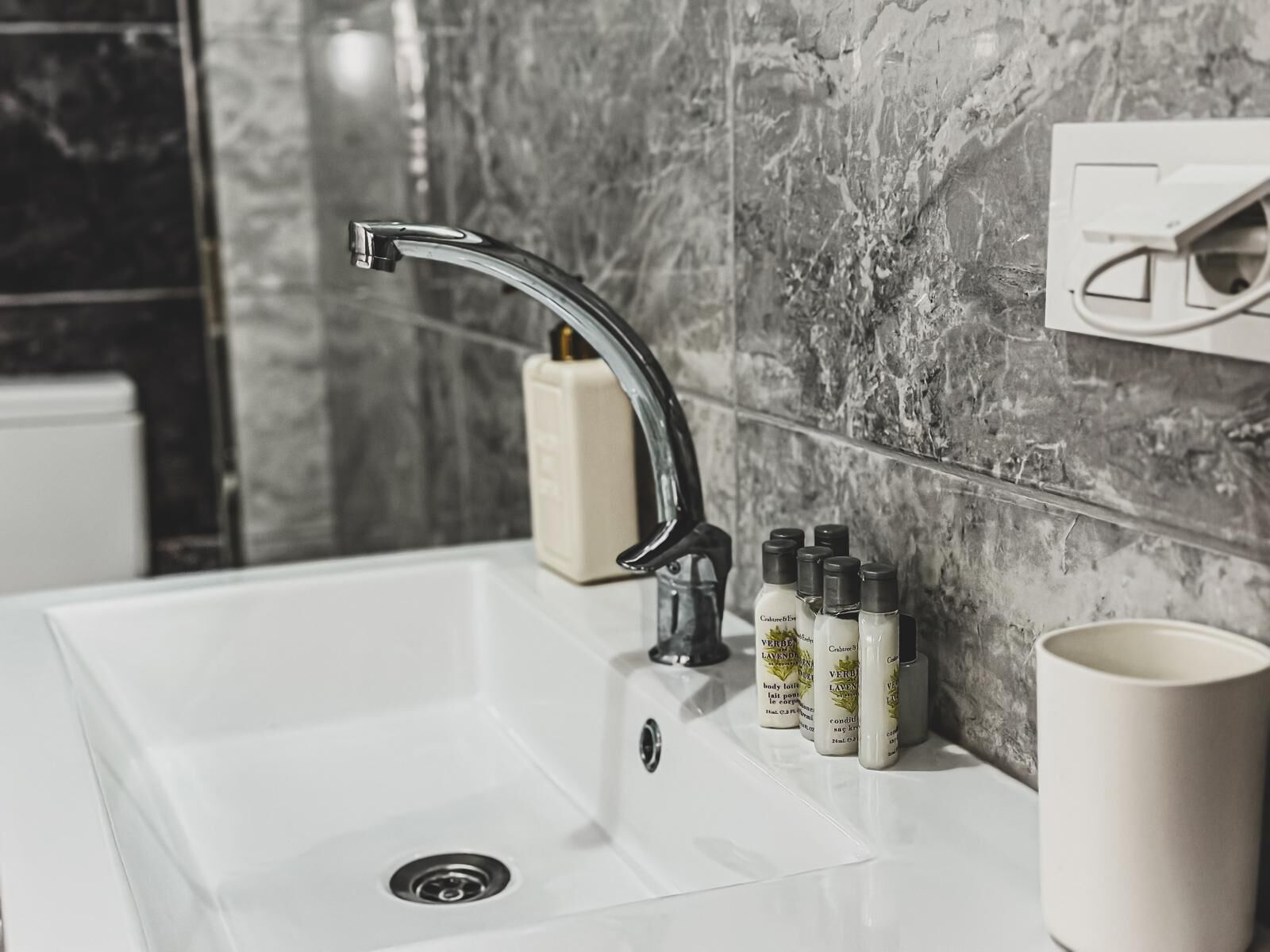 Un baño con lavamanos blanco, un grifo moderno y un salpicadero de mármol. Hay artículos de aseo en el lavamanos y una taza blanca en la encimera.