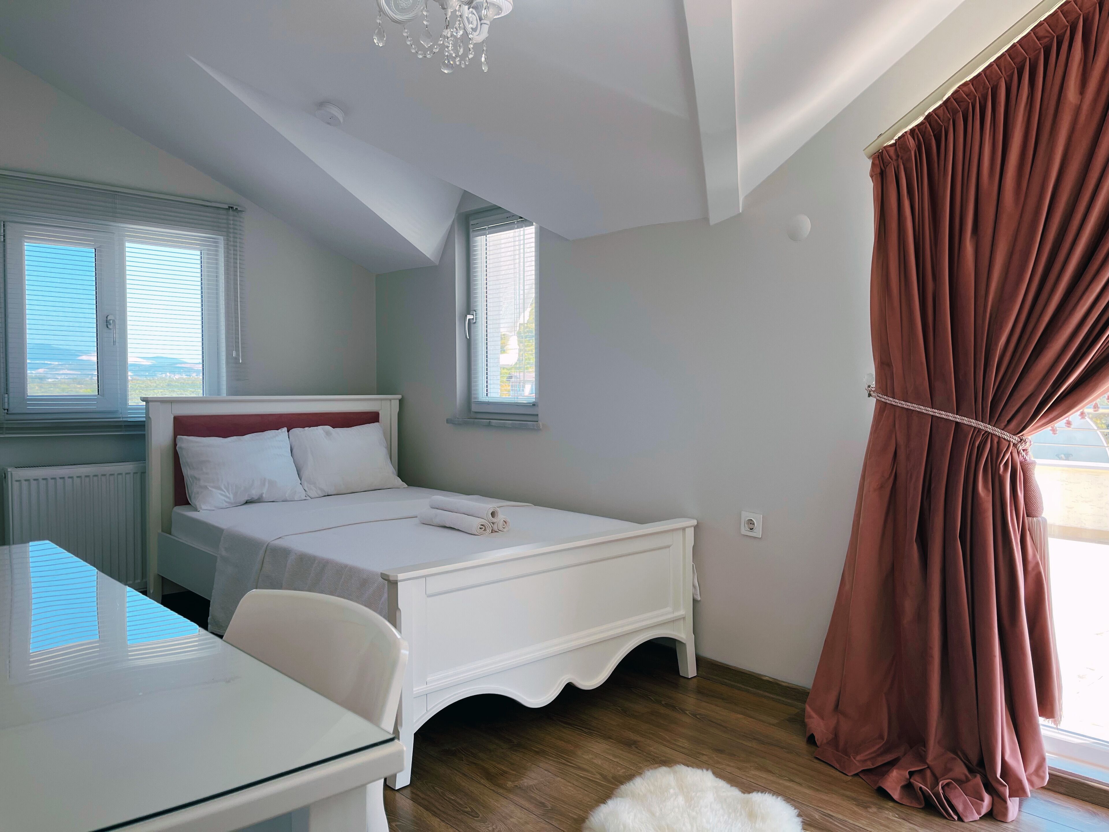A bedroom with a bed, a desk, a chair, a window with blinds, a curtain, and a chandelier.