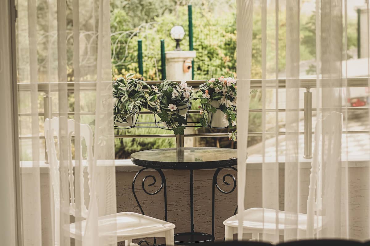 Un balcon agrémenté d’une table et de chaises, entouré de rideaux blancs, avec des plantes en pot et une vue sur la verdure.