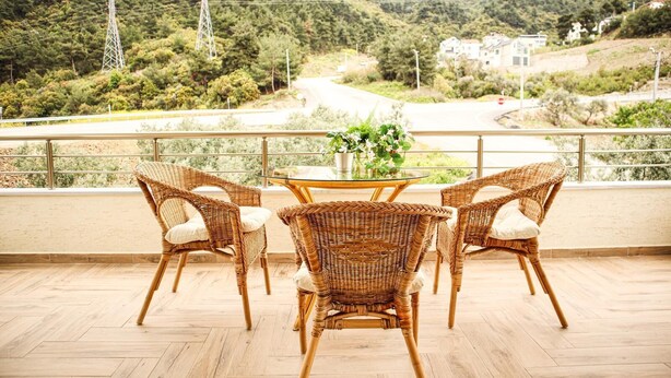 Un balcon avec des fauteuils en osier et une table en verre, donnant sur une route et des collines.