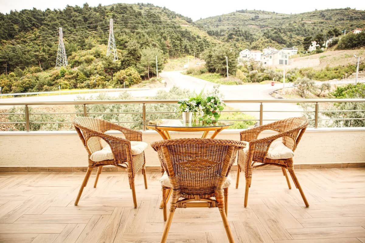 Un balcon avec des fauteuils en osier et une table en verre, donnant sur une route et des collines.