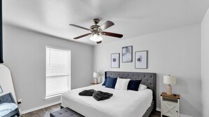A bedroom with a large bed, a ceiling fan, a window with blinds, a bedside table with a lamp, and a small nightstand.