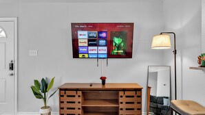 A modern living room with a mounted TV displaying various streaming services, a wooden console table with a vase of flowers, a floor lamp, and a large mirror reflecting the room.