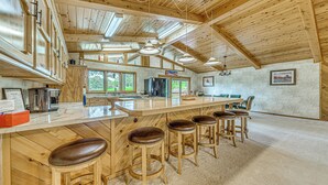 A spacious kitchen with wooden cabinetry, a large island with stools, and a view of the outdoors through large windows. - Hot Tub & Deck: Home 1 Mi to Ruidoso River! (Ruidoso)