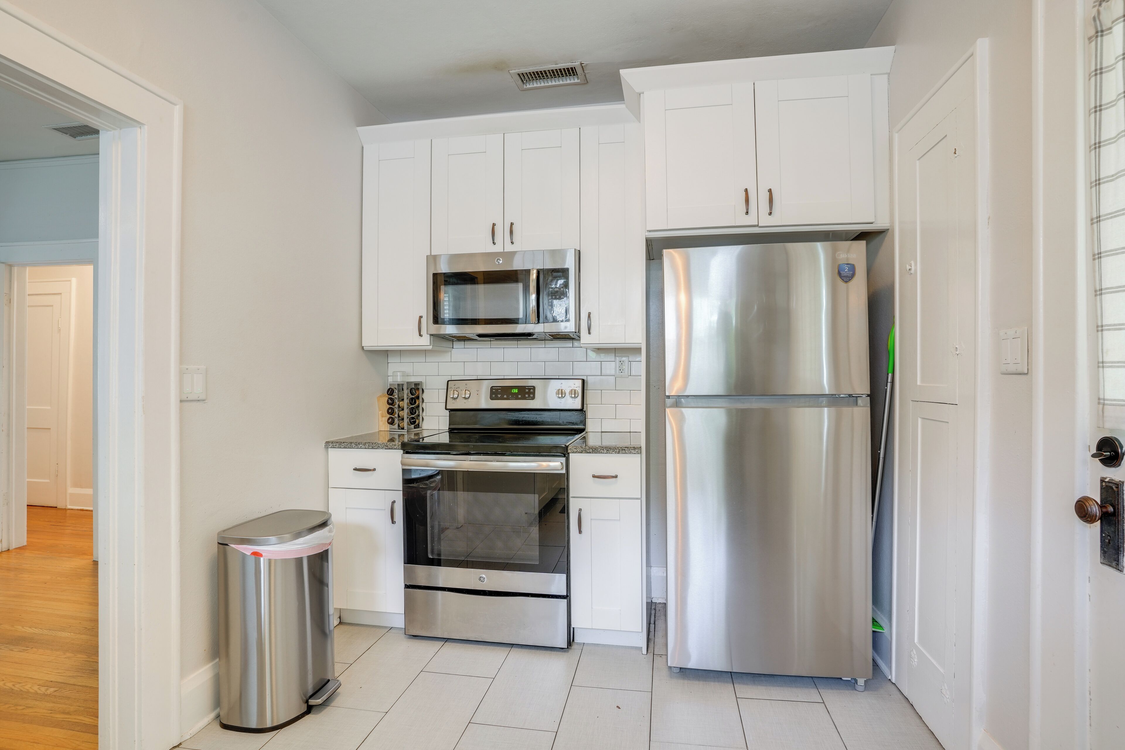 A kitchen with white cabinets, a stainless steel refrigerator, a microwave, an oven, and a trash bin.