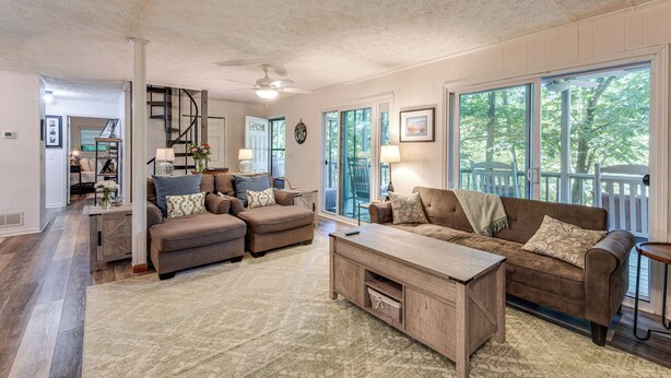 A spacious living room with a large window, a ceiling fan, and a staircase leading to another level. - Hiwassee River Gem w/ Huge Decks: Great for Groups (Hiawassee)