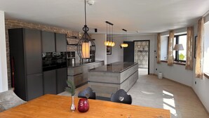 A modern kitchen with dark cabinetry, a central island, pendant lighting, and a dining area with a wooden table. - The Alpine No 7 (Reith bei Seefeld)