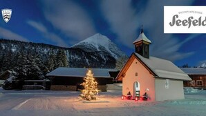 A snow-covered church with a lit Christmas tree in front, surrounded by buildings and a mountain in the background. - The Alpine No 7 (Reith bei Seefeld)