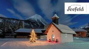 A snow-covered church with a lit Christmas tree in front, surrounded by buildings and a mountain in the background. - The Alpine No 7 (Reith bei Seefeld)
