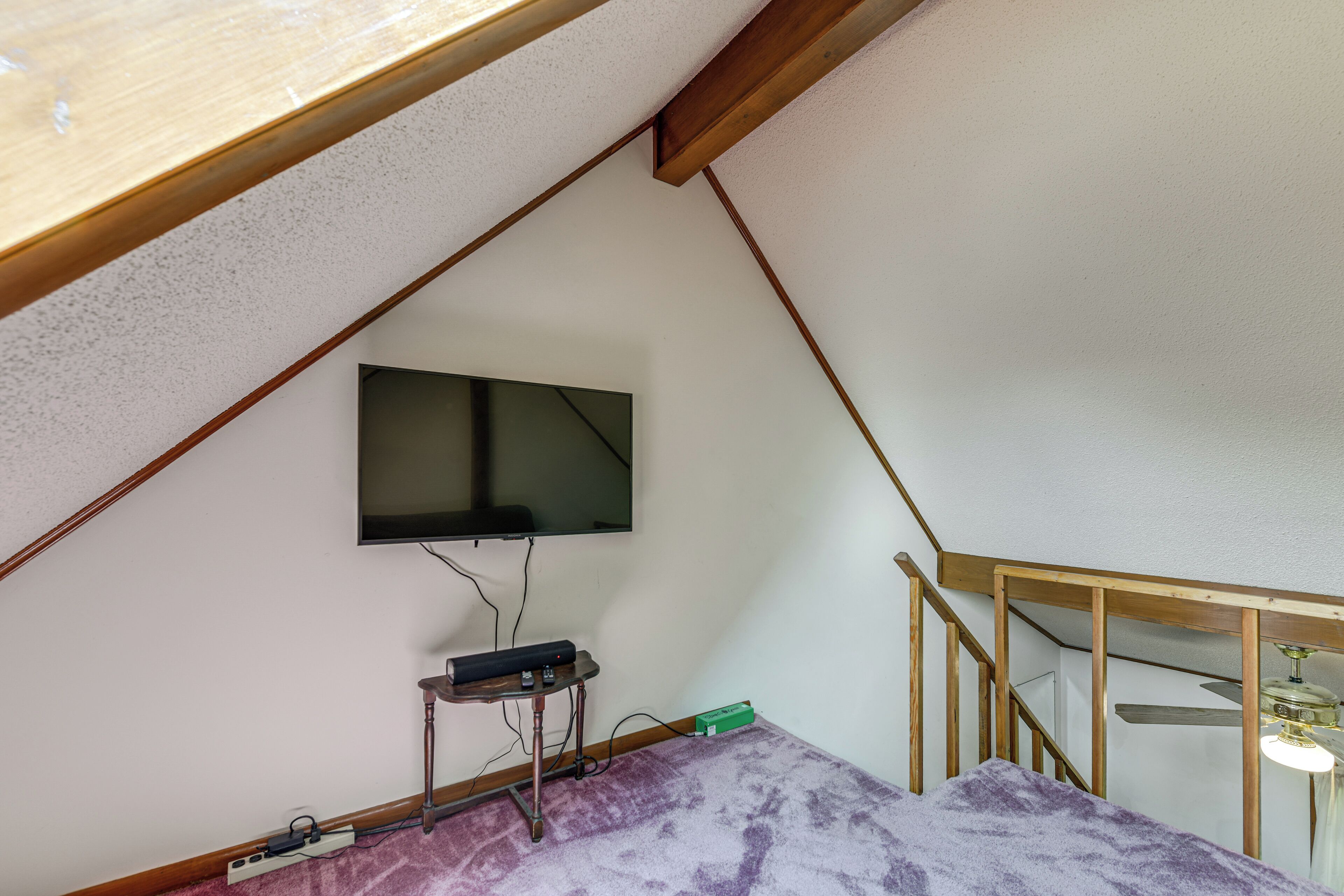 A room with a sloped ceiling, a mounted television, a small table with electronic devices, and a staircase with a wooden railing.