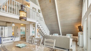 A spacious loft with a high ceiling, a dining area with a wooden table and chairs, a kitchen with white cabinetry, and a staircase leading to an upper level.