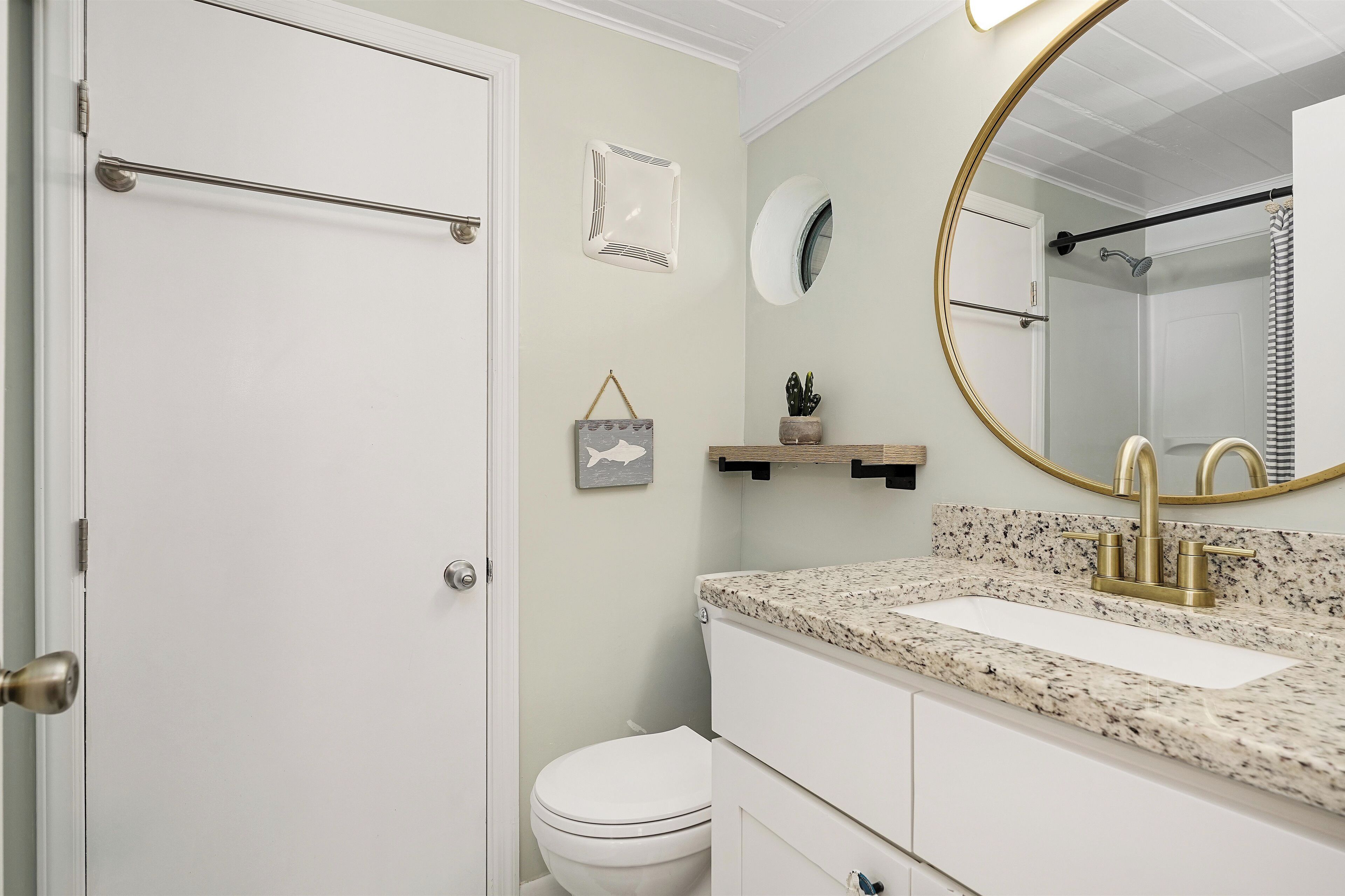Une salle de bain avec des toilettes blanches, un lavabo doté d’un plan de travail en granit, un robinet doré, un miroir rond et un rideau de douche.