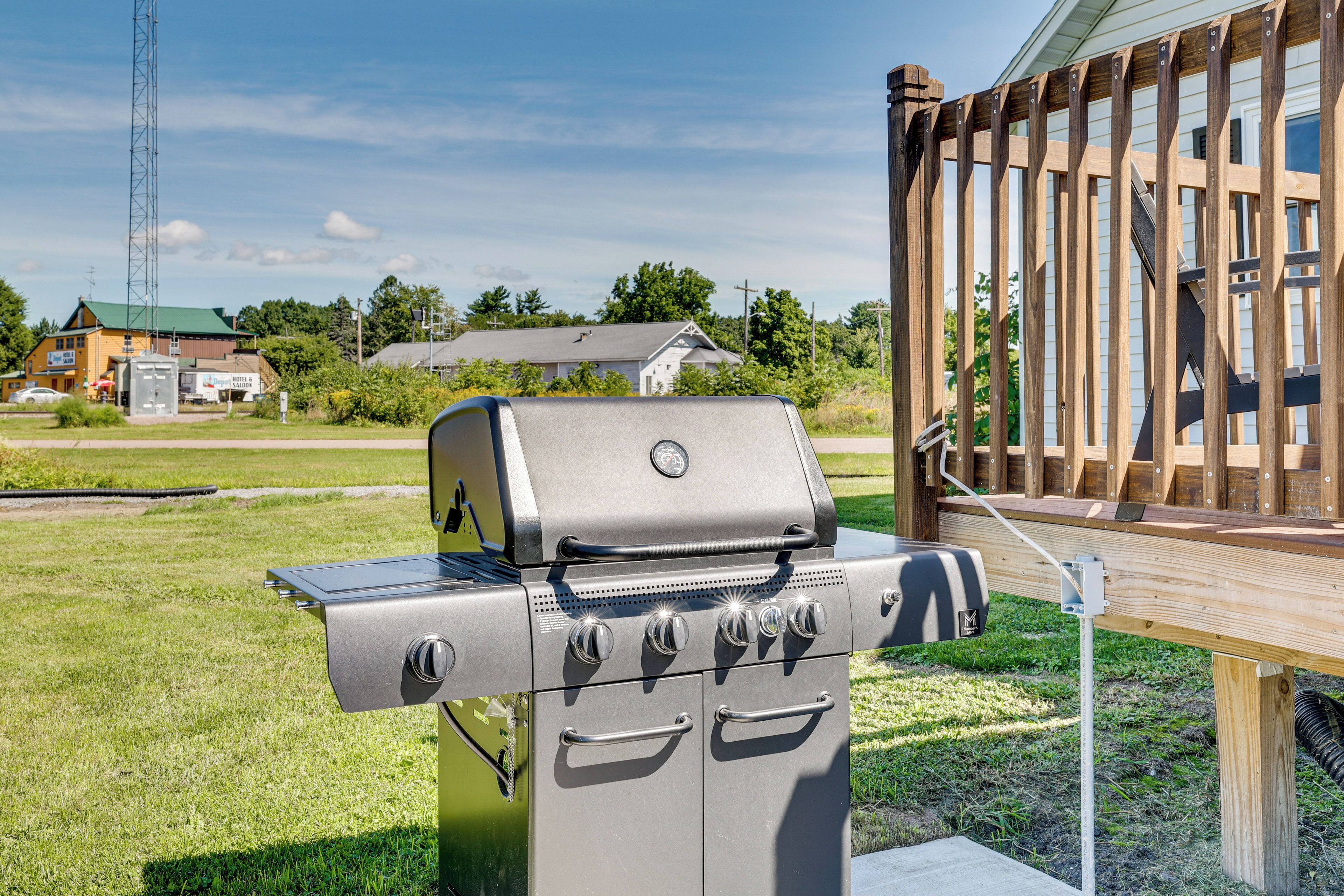 A stainless steel outdoor grill with four burners and a side shelf, set against a grassy lawn with a wooden deck and a house in the background.