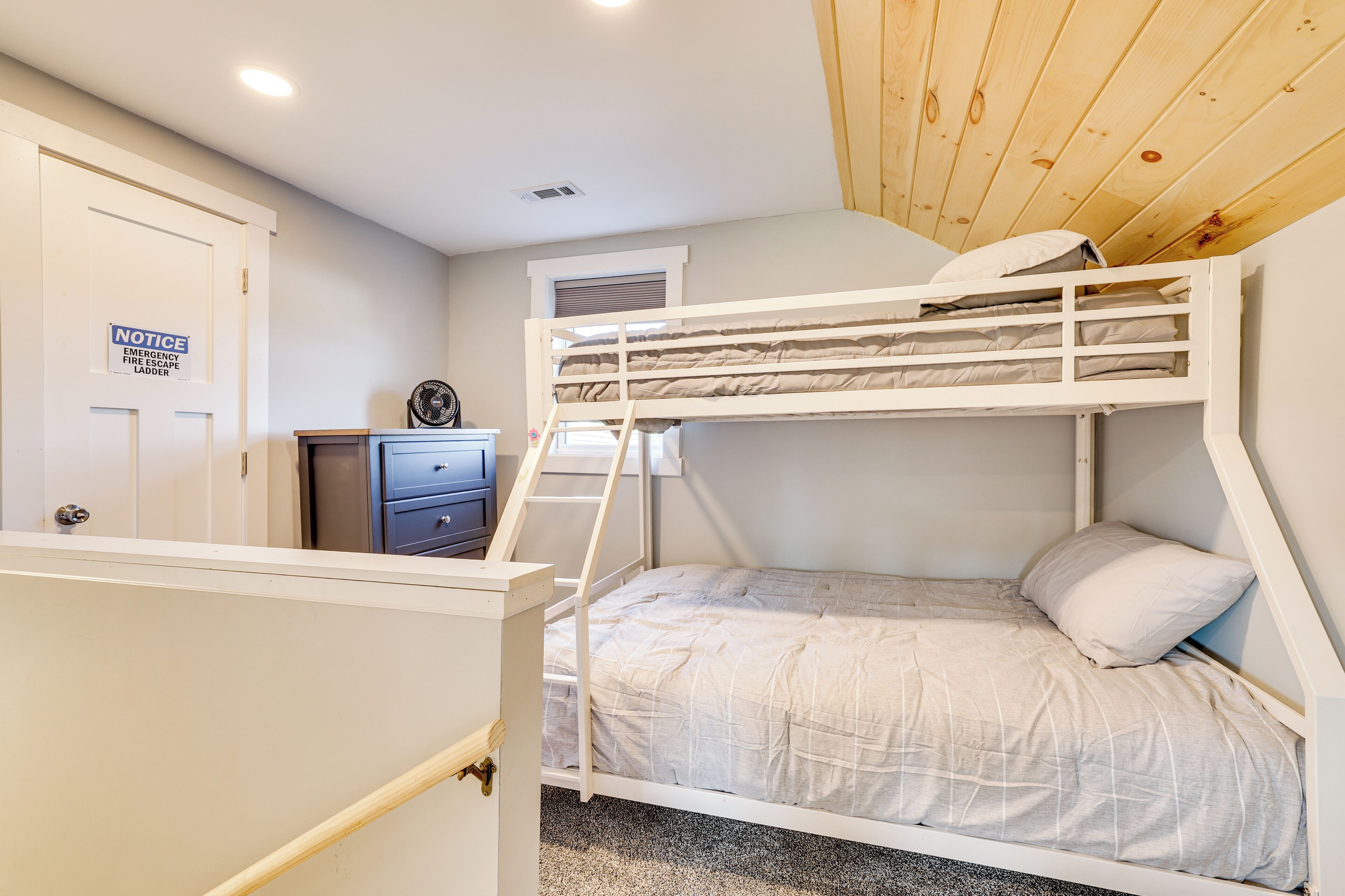 Een stapelbed met een ladder, een bureau met lades, een deur met een bordje en een raam met jaloezieën.