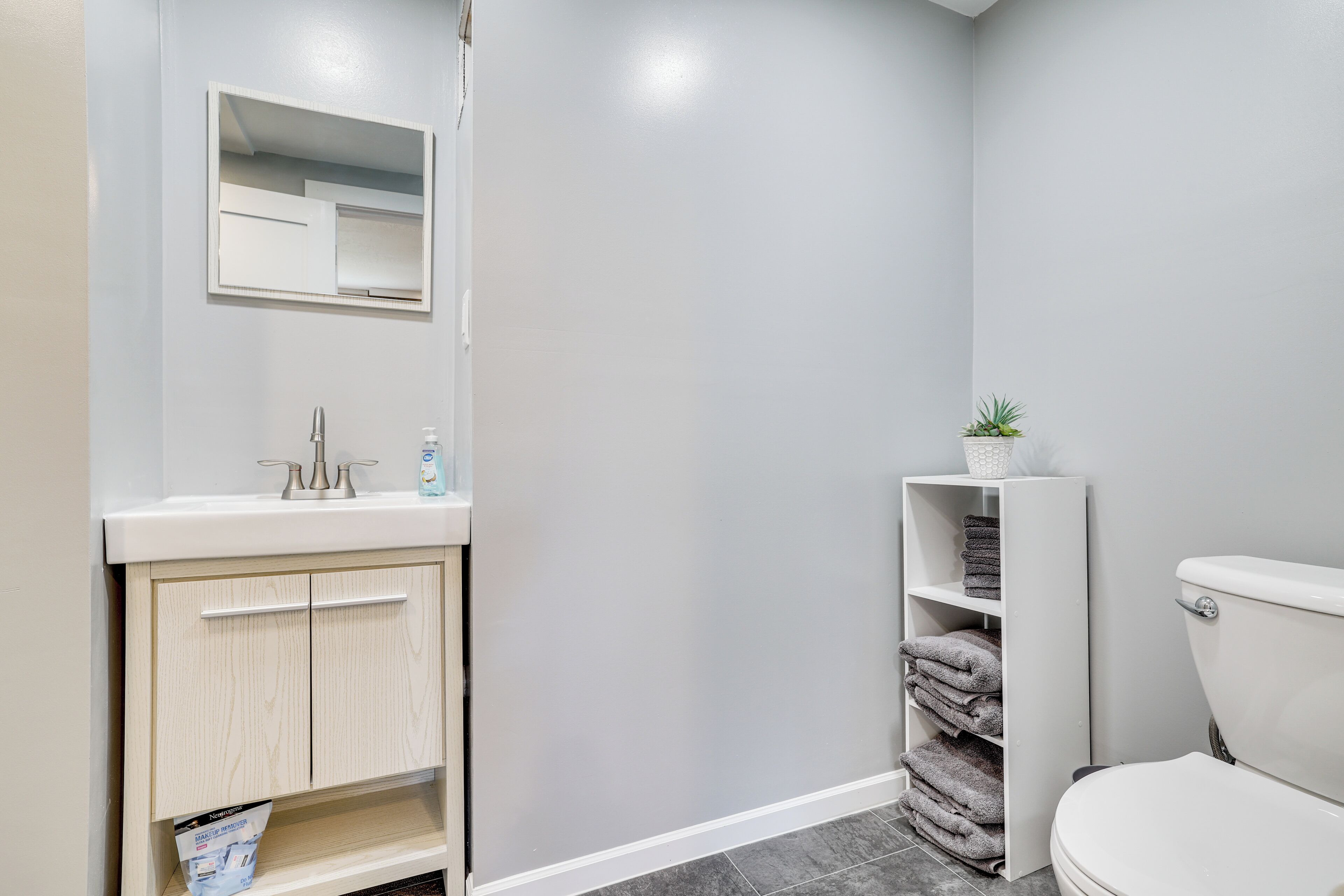 A bathroom with a white sink, mirror, and toilet. There is a shelf with folded towels and a small plant.
