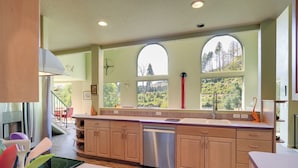 A modern kitchen with a dishwasher, wooden cabinets, and large windows offering a view of trees. - On Mckenzie River! Home w/ Scenic Sunroom (Vida)