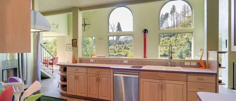 A modern kitchen with a dishwasher, wooden cabinets, and large windows offering a view of trees.