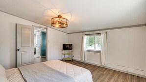 Un dormitorio con cama, televisor sobre un mueble, ventana con cortinas y puerta que da a otra habitación.