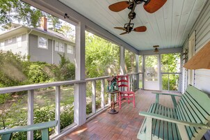 A covered porch with a bench and a red chair, a birdhouse, and a view of a house and greenery.