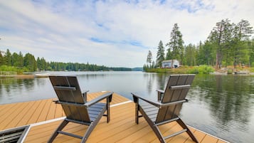 A wooden deck with two chairs facing a calm lake, surrounded by trees and a small cabin in the distance.