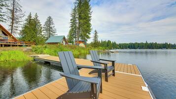A wooden dock with two Adirondack chairs, a small cabin, and a forested shoreline.