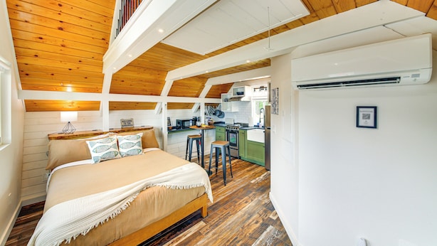 Een slaapkamer met een houten plafond, een bed met een beige deken en een kitchenette op de achtergrond.