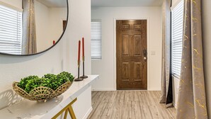 A hallway with a wooden door, a white wall with a mirror, and a white console table with a plant and candles. - Modern Comforts + Fenced Yard: Princeton Retreat! (Princeton)