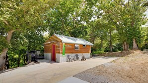 Une petite cabane en bois avec un toit en métal, une porte rouge et une petite fenêtre. Devant la cabane, il y a une zone pavée, et des arbres entourent les lieux.