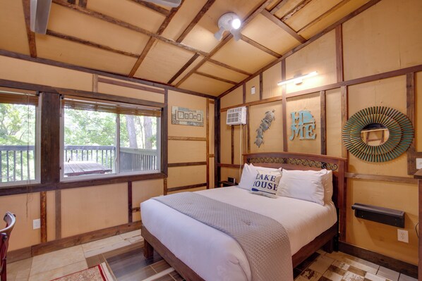 Un dormitorio con techo de madera, una cama con ropa blanca, un ventanal con vistas a árboles y una pared decorada.
