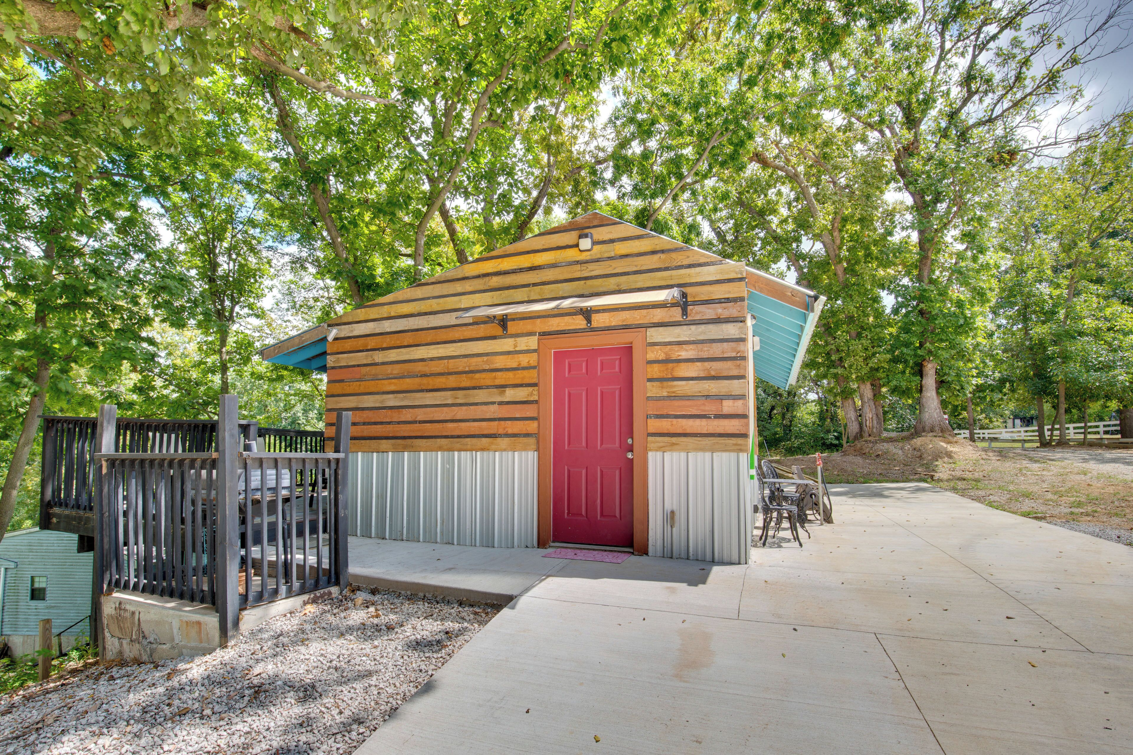 A small wooden cabin with a red door, surrounded by trees and a concrete patio.